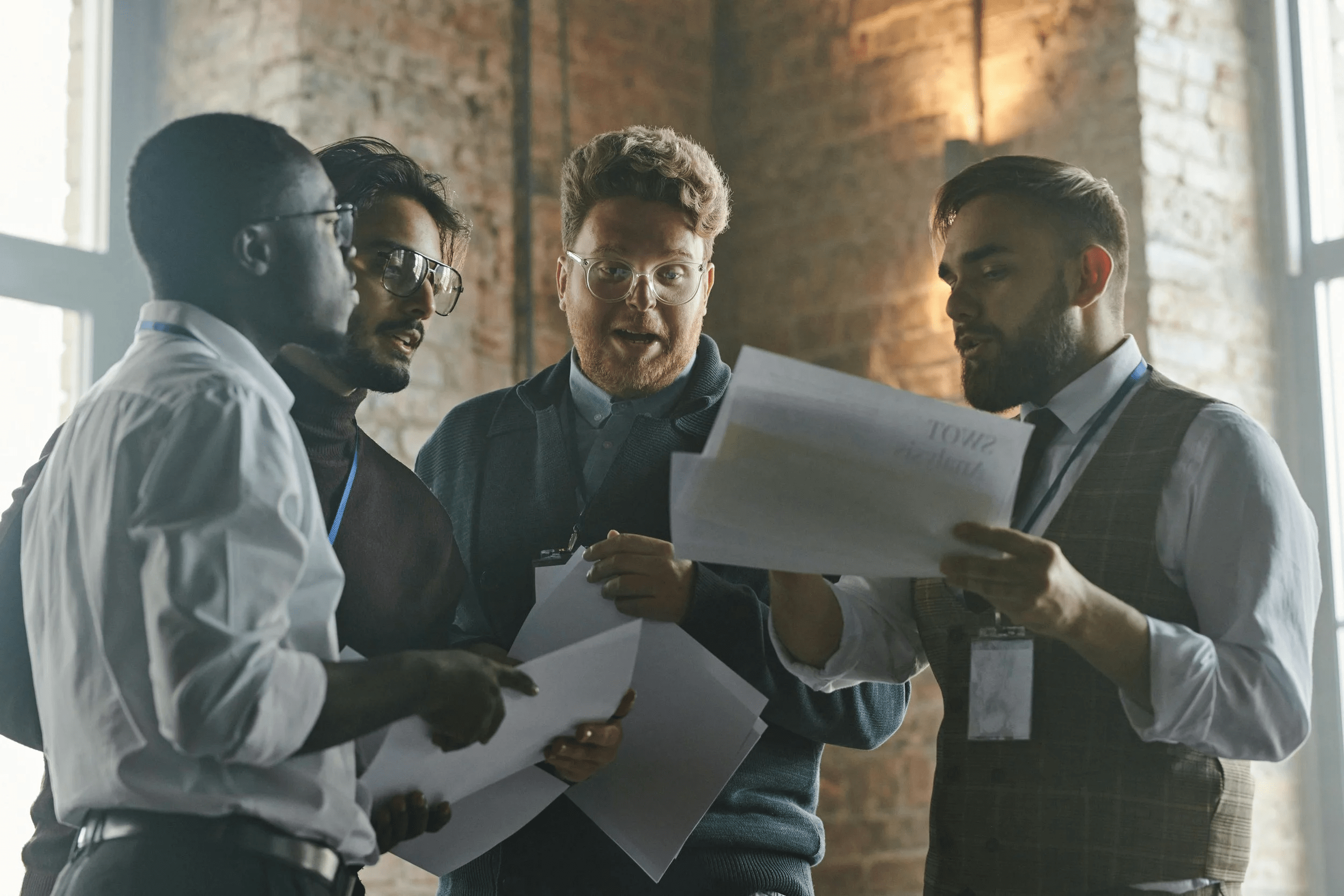 Four men are engaged in a discussion while looking at papers in a well-lit room with brick walls.