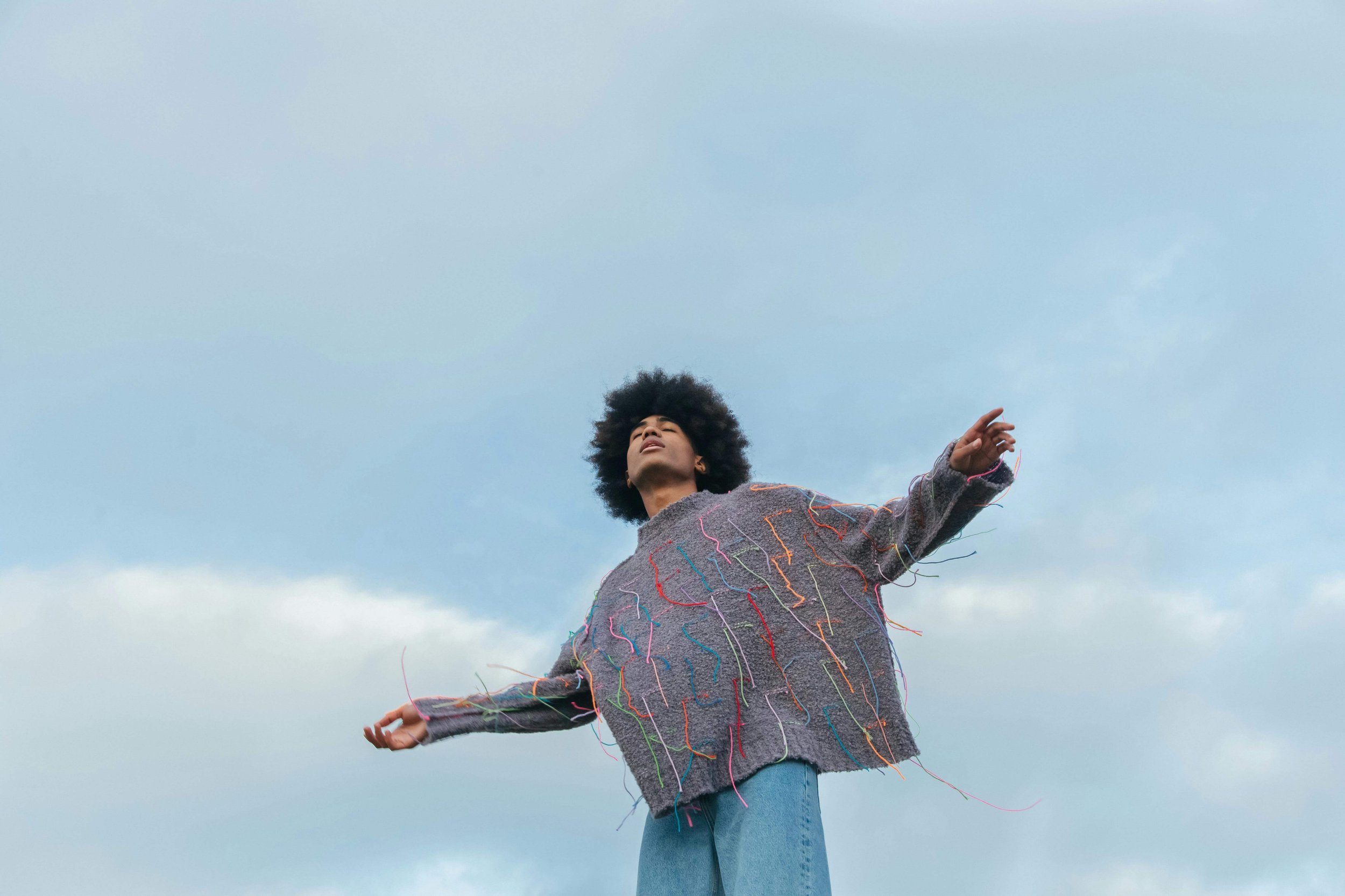 A person with curly black hair stands outdoors against a cloudy sky, wearing a gray oversized sweater with colorful threads and blue jeans, holding arms outstretched with eyes closed.