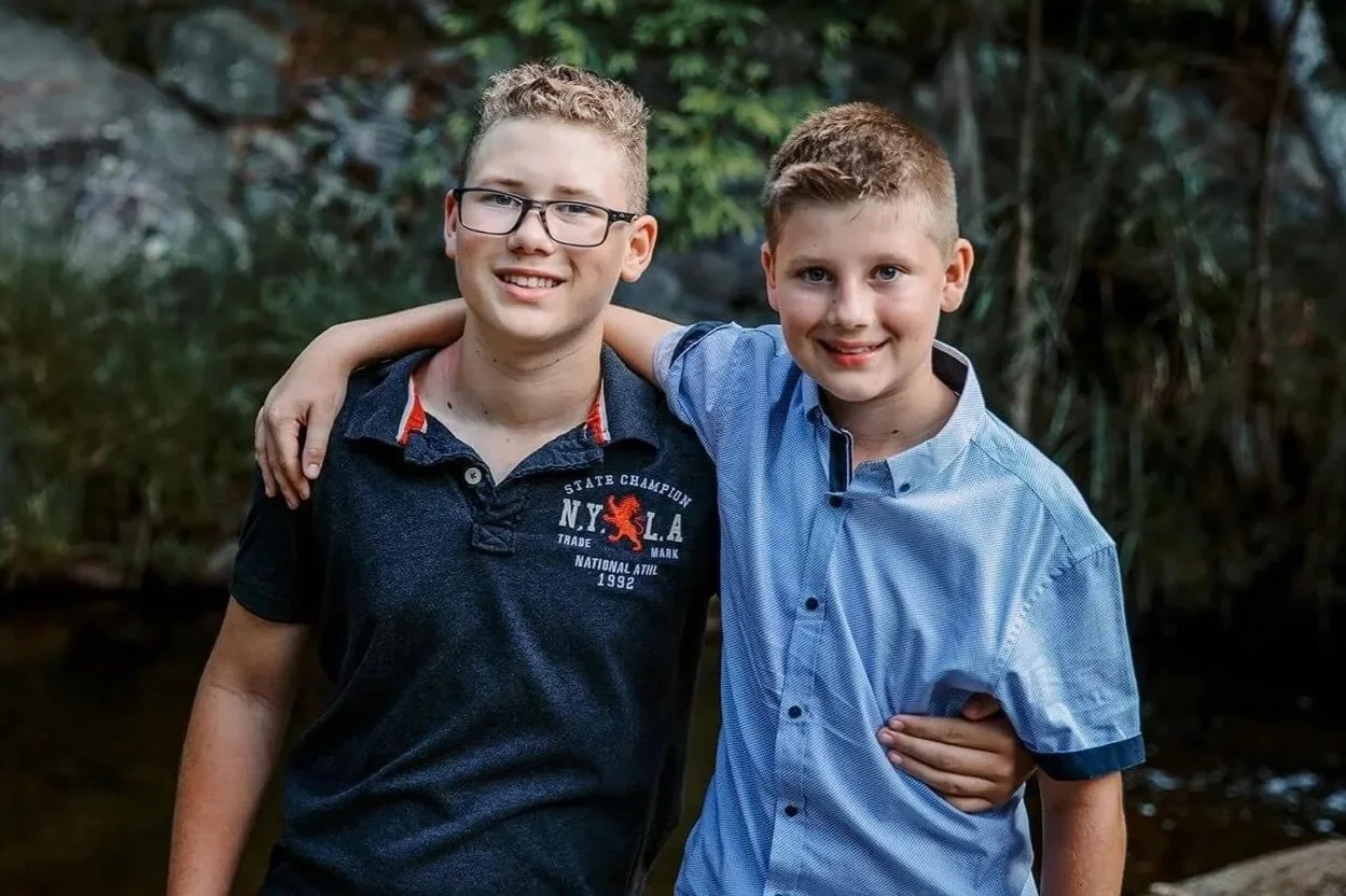 Two young boys with light skin, smiling with arms around each other, standing outdoors with trees and water in the background.