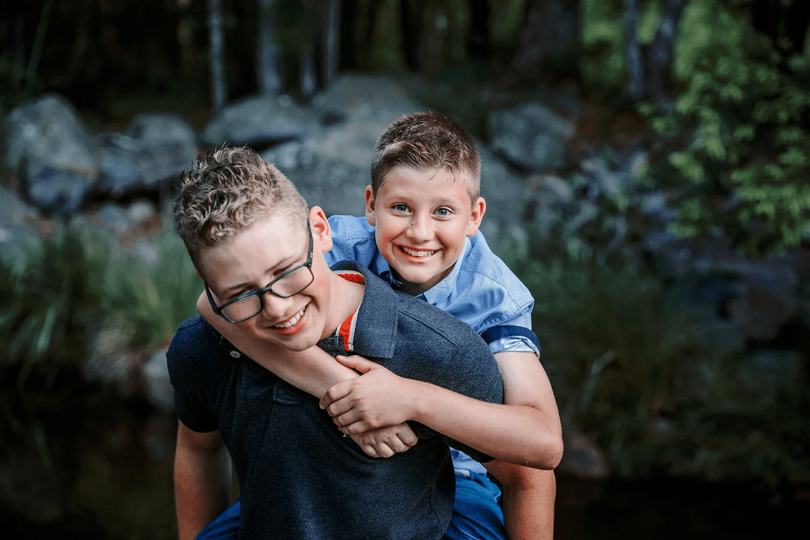 Two boys smiling and playing outdoors, one giving a piggyback ride on the other's back, set in a natural, wooded area.