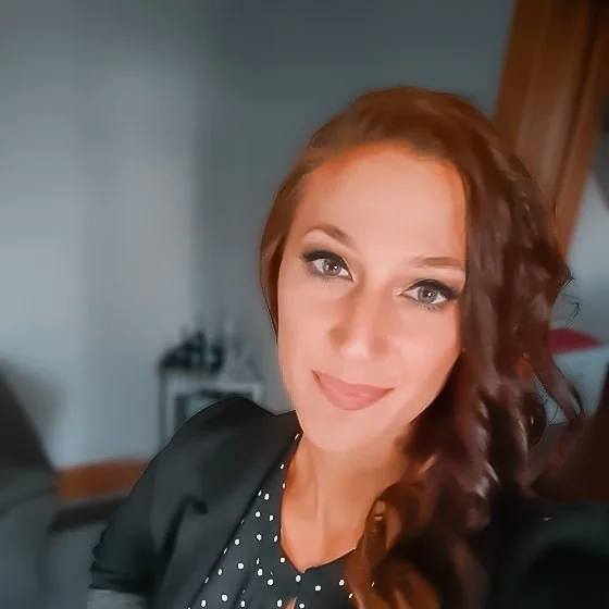 A woman with long, wavy red hair and light makeup smiling at the camera, wearing a black top with white polka dots, in a room with a blurred background.