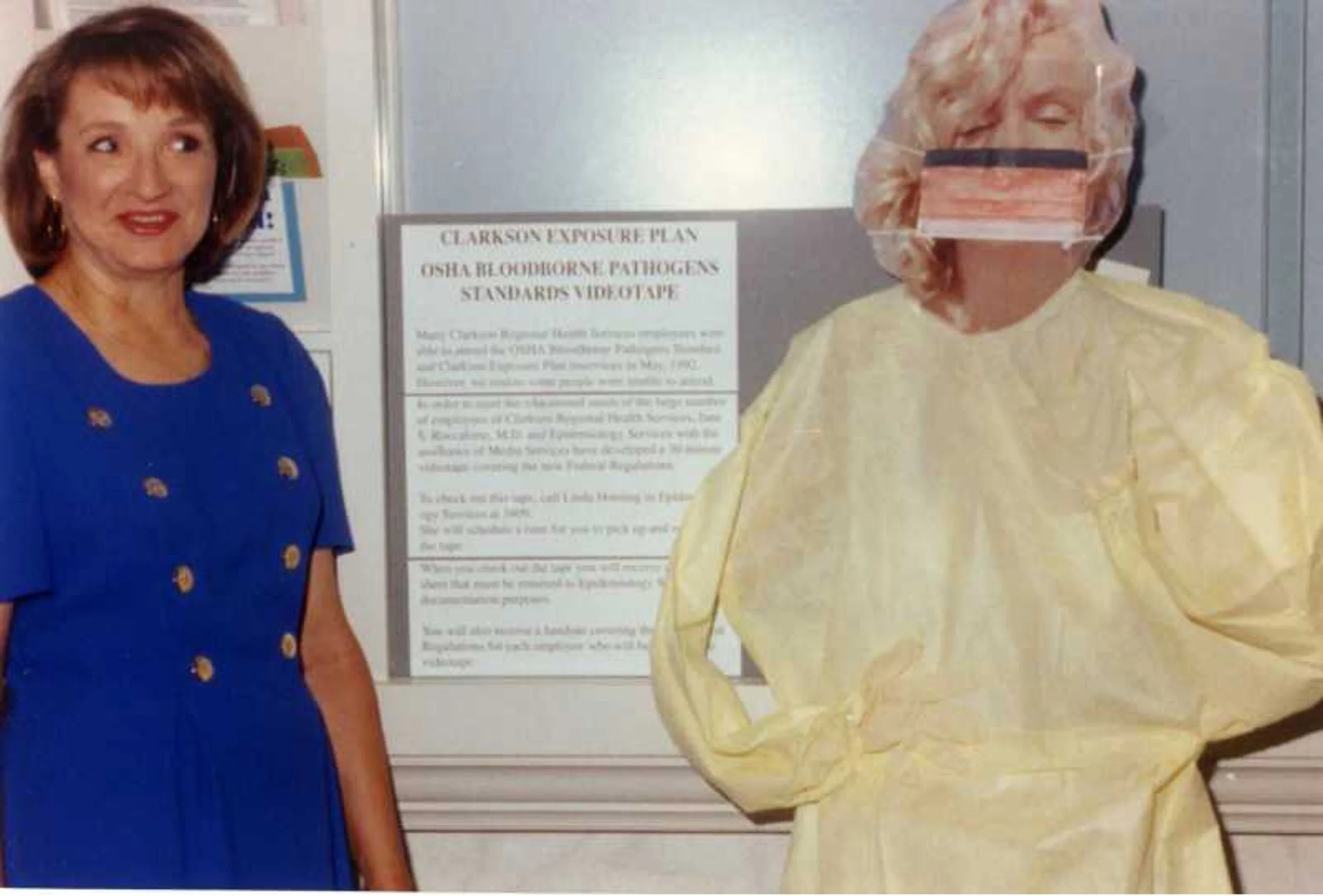 Two women standing next to each other, one in a blue dress and the other in yellow protective clothing with a face mask, in front of a sign about OSHA pathogens standards.