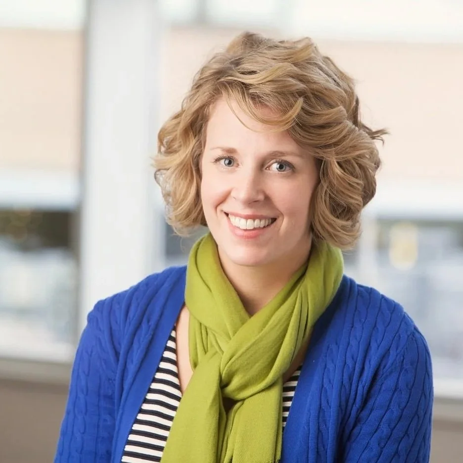 A smiling woman with short, curly blonde hair, wearing a green scarf, a blue cardigan, and a black and white striped top, standing indoors near a window.
