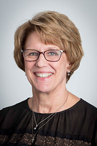 A smiling older woman with short, light brown hair, wearing glasses and a black top, against a plain background.