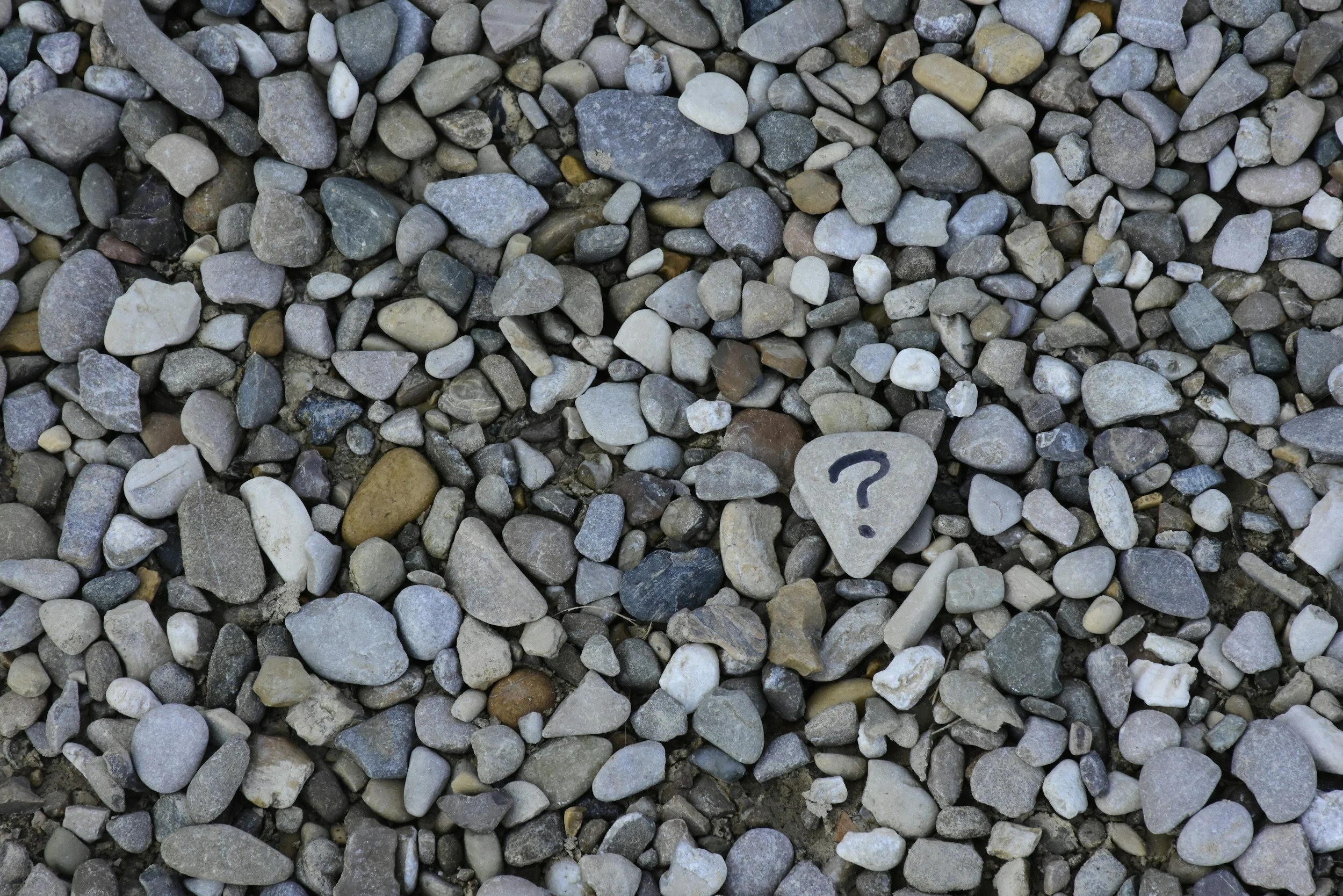A collection of small, multicolored pebbles on the ground with a single pebble that has a black question mark drawn on it.