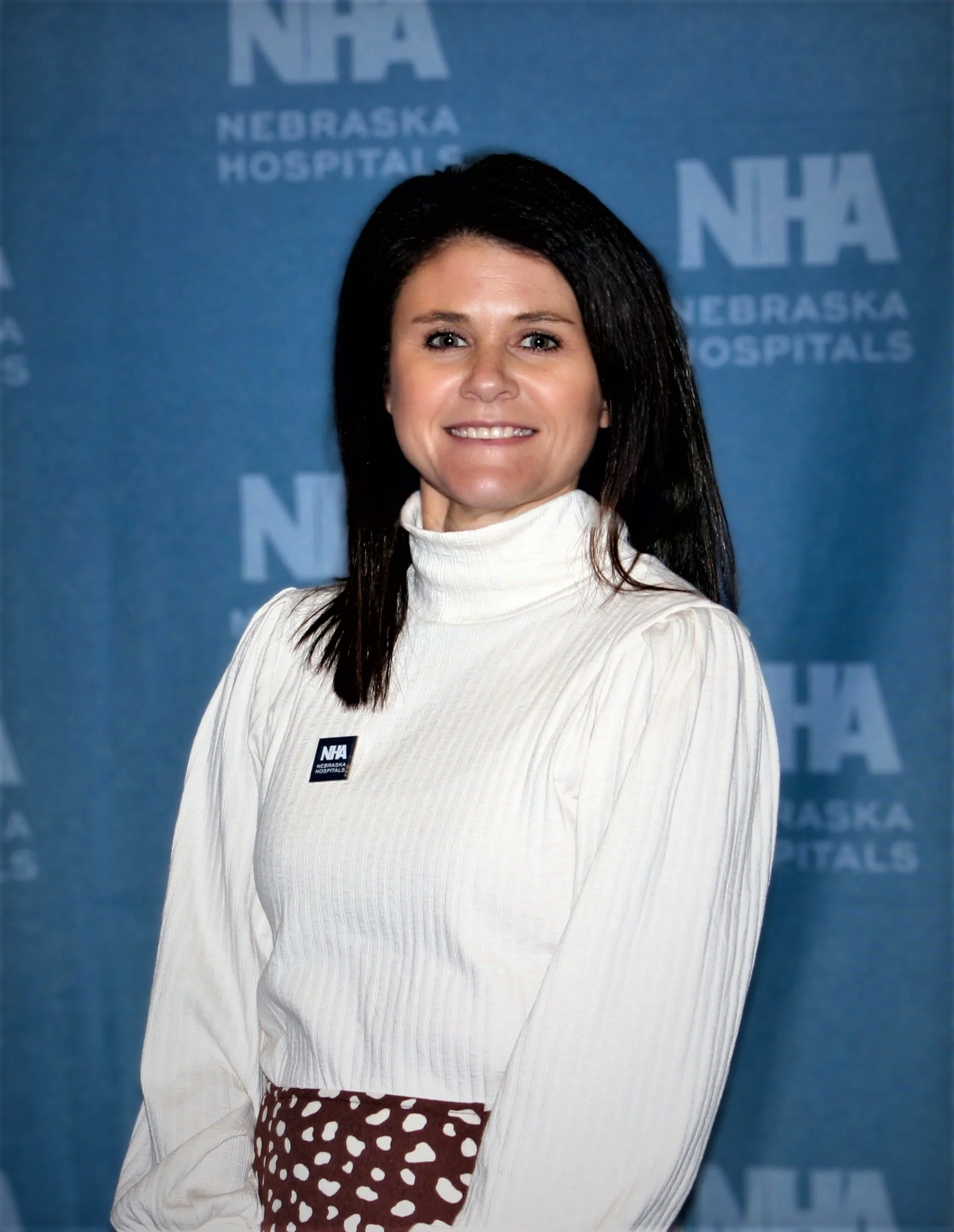 A woman with dark hair wearing a white turtleneck sweater and a patterned skirt, standing in front of a blue Nebraska Hospitals backdrop.