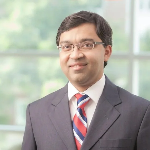 Professional man in a suit and tie, wearing glasses, smiling in front of a blurred office background.
