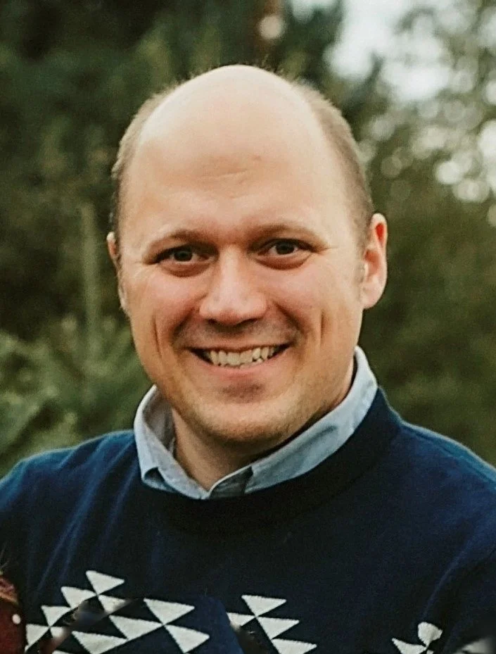 Smiling man in a navy sweater and collared shirt outdoors with blurred trees in the background.