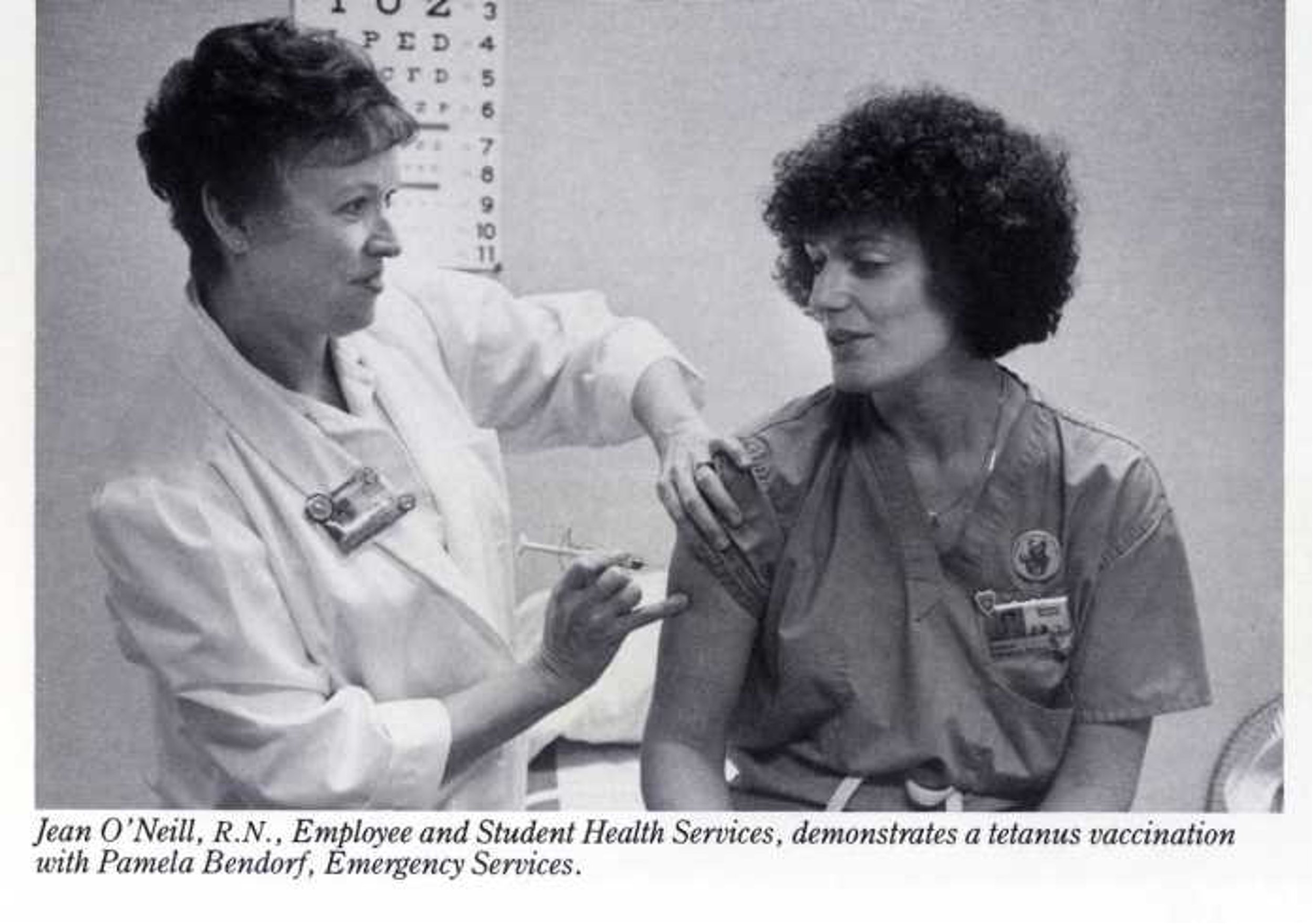 A nurse administering a tetanus vaccination to a woman in medical uniform. The woman is seated with her sleeve rolled up, while the nurse is focused on giving the shot. There is a height measurement chart on the wall behind them.