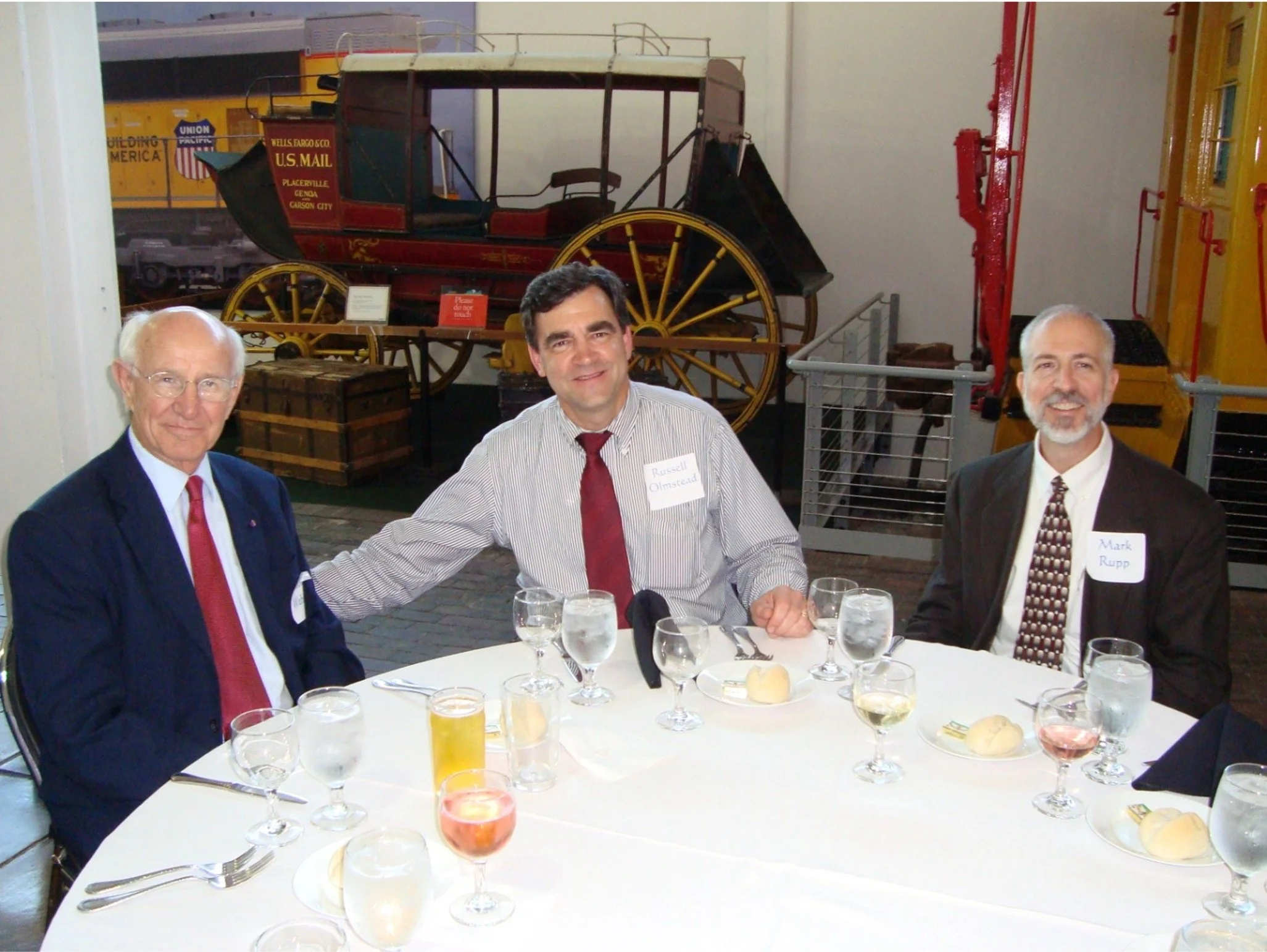 Three men in suits seated at a round table set for a meal, with a vintage horse-drawn carriage displayed behind them in a museum setting.