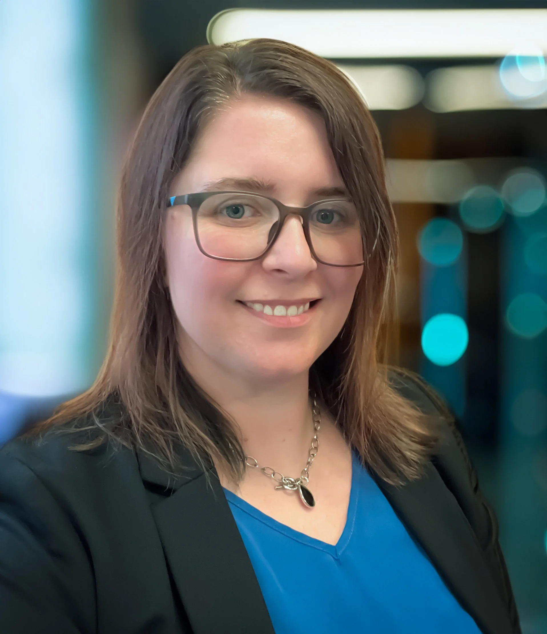 A woman with shoulder-length brown hair, wearing glasses, a blue top, a black blazer, and a silver necklace, smiling at the camera with a blurred background of lights.