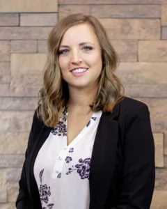 A woman with shoulder-length wavy hair, wearing a black blazer over a white blouse with floral patterns, standing against a brick wall background.