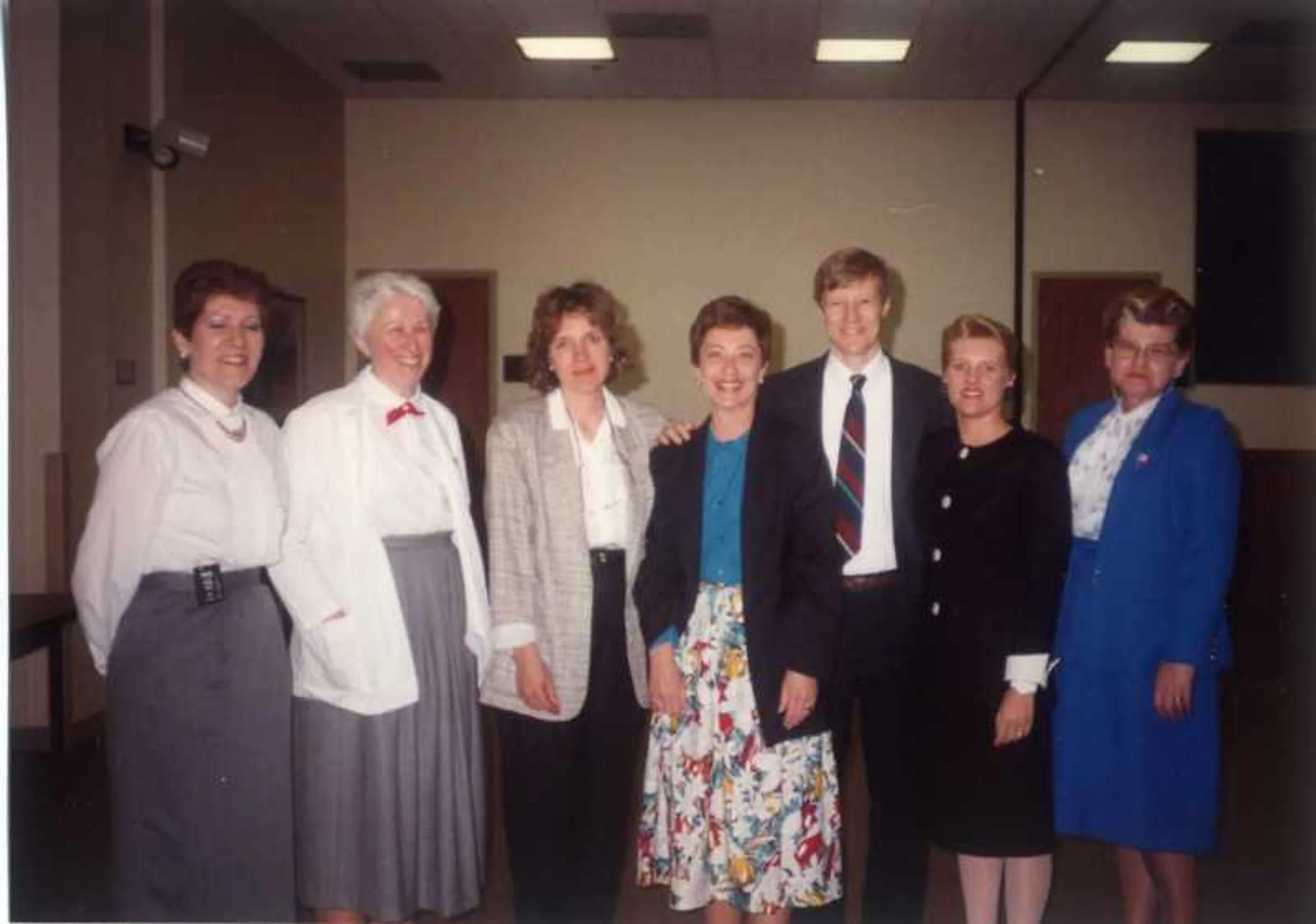 Group of seven women and one man standing side by side indoors, all smiling, dressed in professional attire, with a plain beige wall and ceiling lights in the background.
