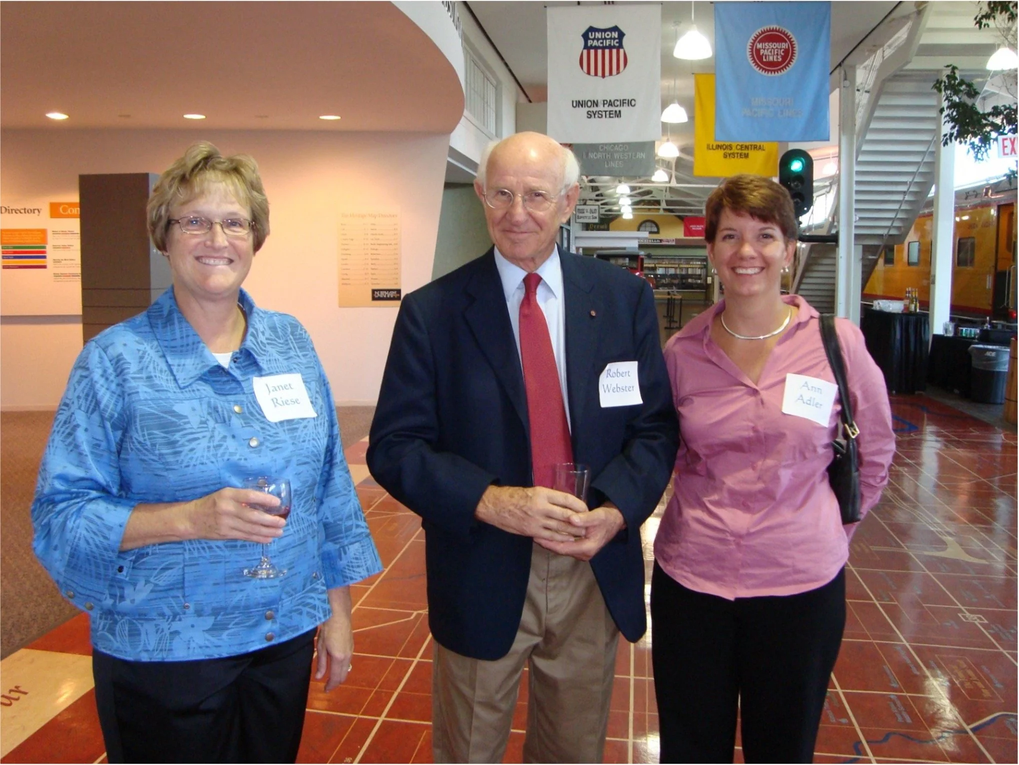 Three people standing inside a building, all smiling and holding drinks. The woman on the left is wearing a blue patterned blouse and glasses, with a name tag that says Janet Riese. The man in the middle is dressed in a suit with a red tie and has a 