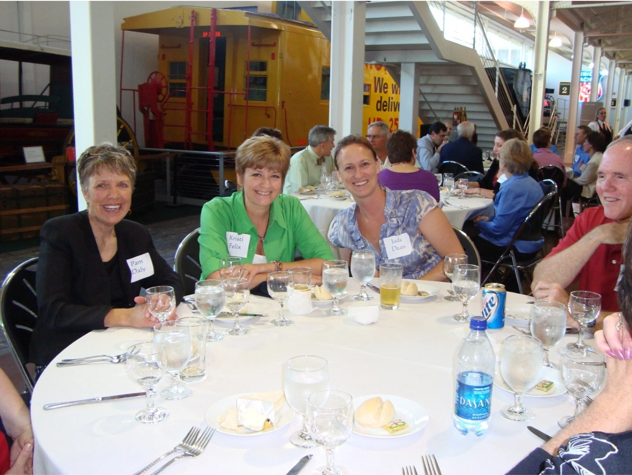 Group of people sitting around a dining table in a large indoor space, with a yellow train car visible in the background.