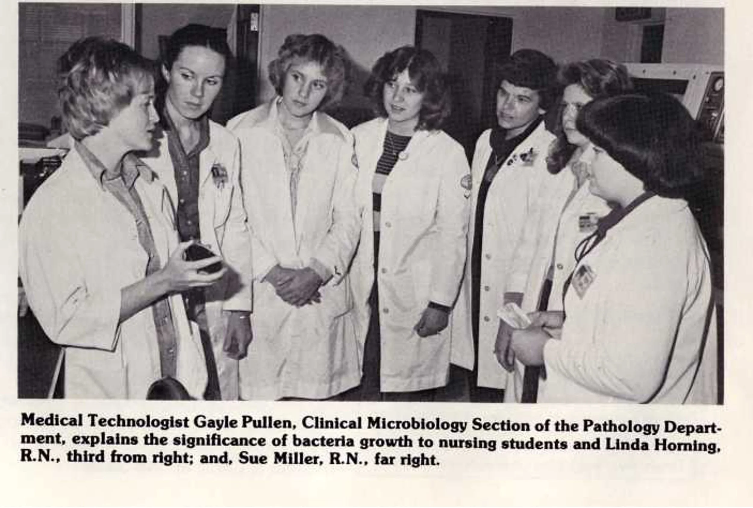 A group of seven women in white lab coats gathered around a woman holding a piece of lab equipment, discussing bacteria growth. They are in a laboratory setting, likely a microbiology or medical research facility.