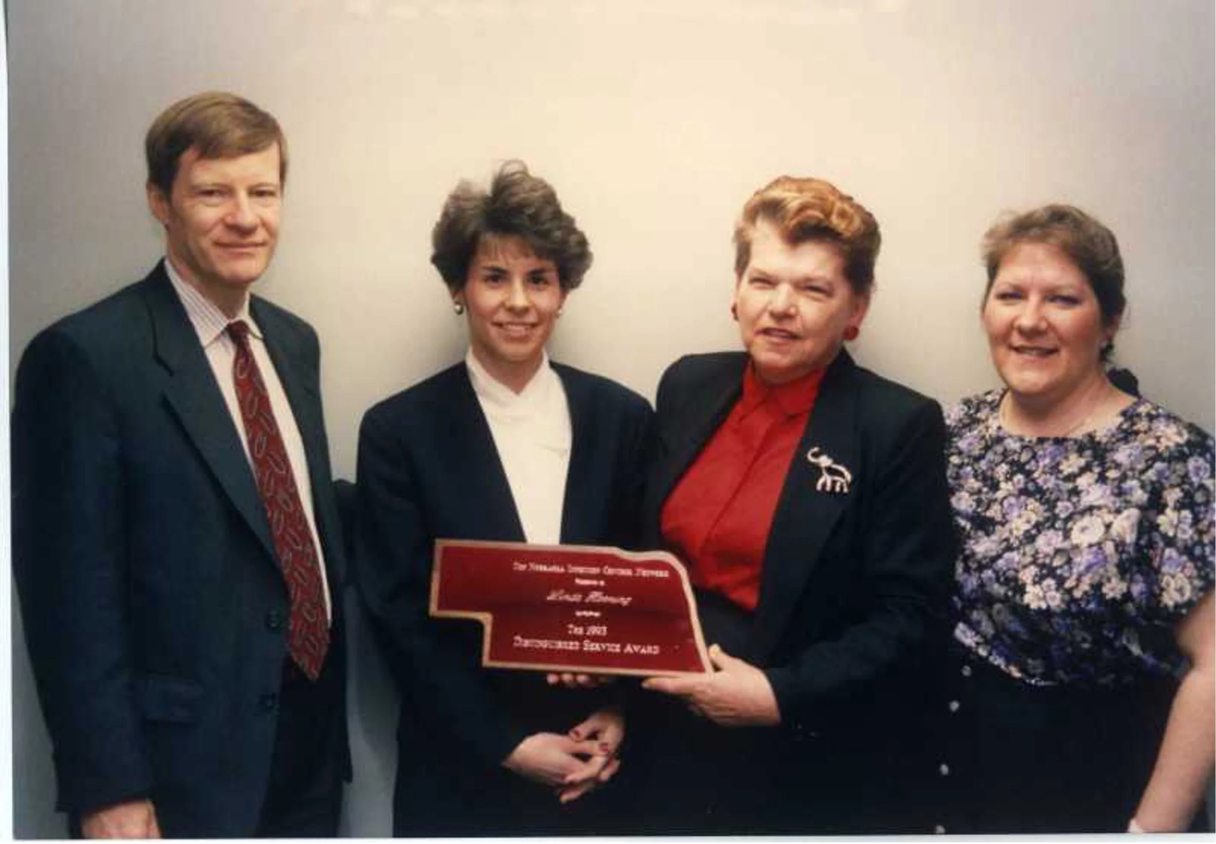 Four women and one man standing together, with the woman second from the right holding an award plaque that reads 'The National Junior Honor Society Award, 1994, Outstanding Service Award.' They are dressed in business attire, smiling, in front of a 