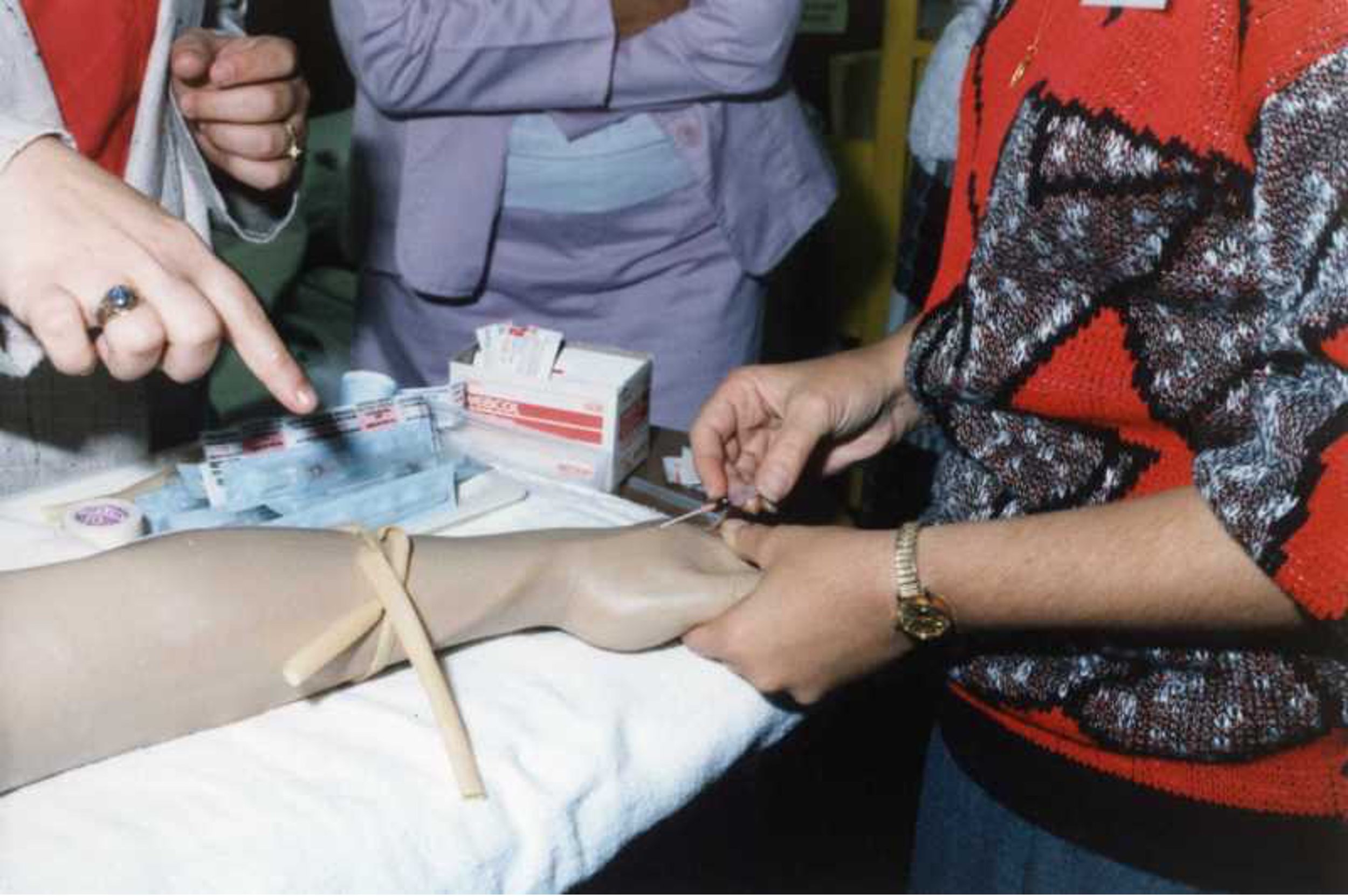 Person receiving an intravenous injection while three people assist. Medical supplies on the table.