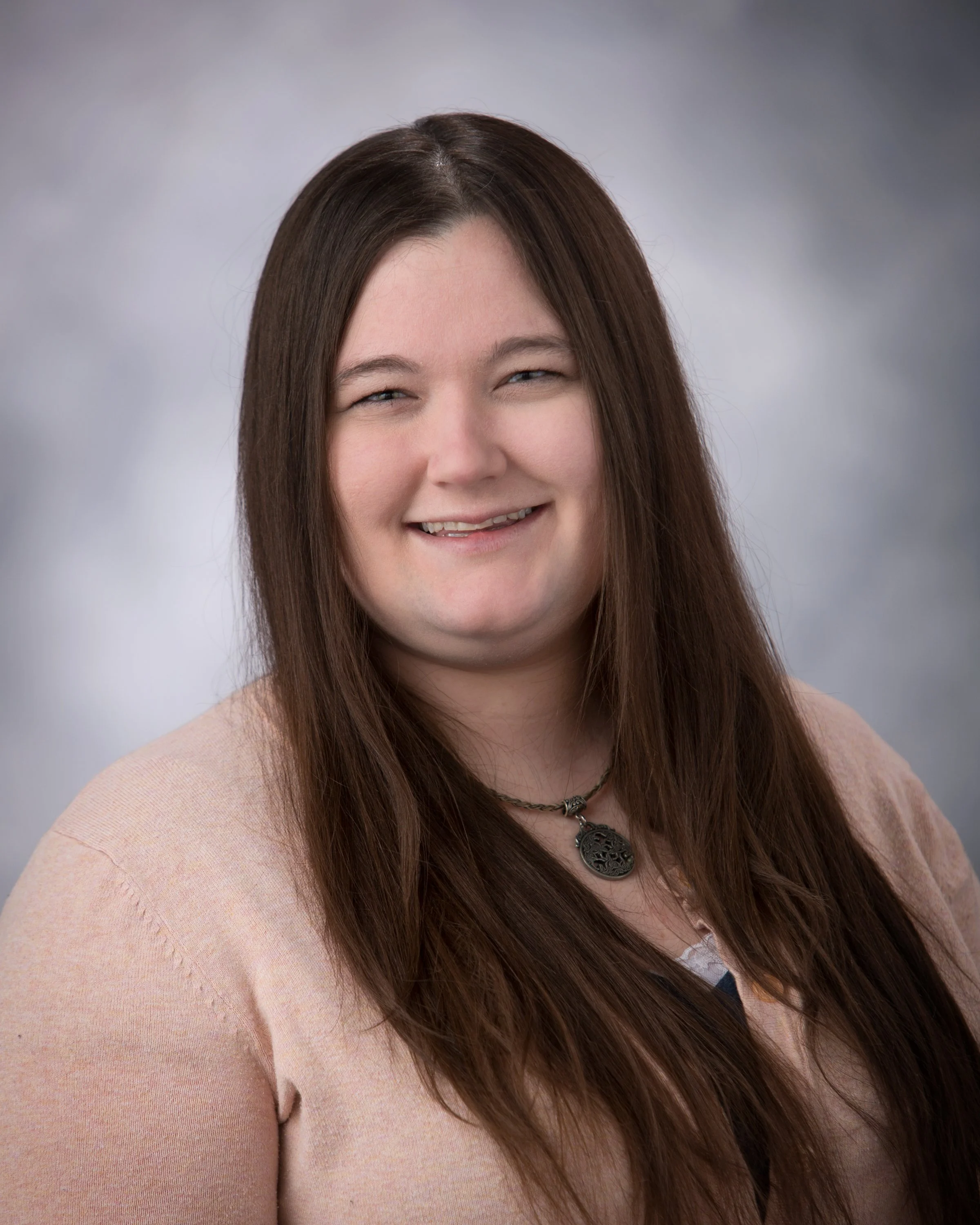 A woman with long brown hair, smiling, wearing a pink sweater and a necklace, with a gray cloudy background.