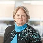 An elderly woman with light-colored hair, wearing a black jacket with floral embroidery and a blue scarf, smiling in an indoor setting.