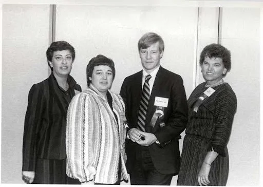 Four women and one man dressed in formal clothing, standing together at an indoor event, with some wearing ribbons or badges.