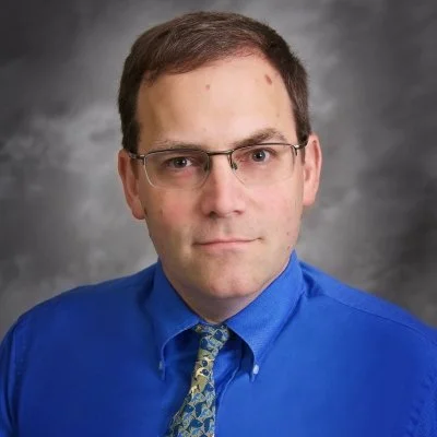 Portrait of a man wearing glasses, a blue shirt, and a patterned tie against a gray background.