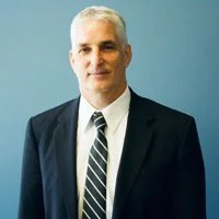 Portrait of a middle-aged man wearing a dark suit, white shirt, and striped tie, standing against a blue background.
