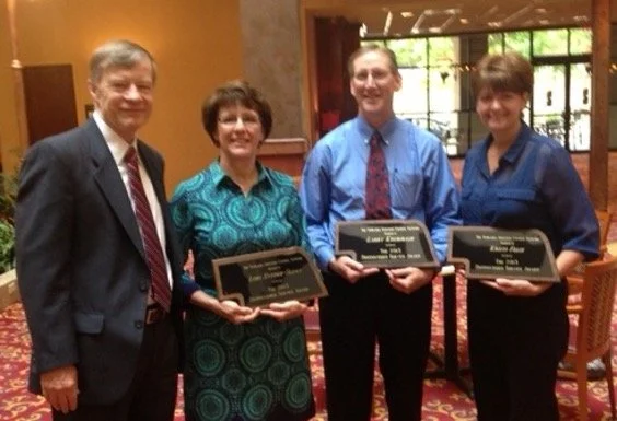 Four people standing in a room, three of them holding plaques, posing for a photo.
