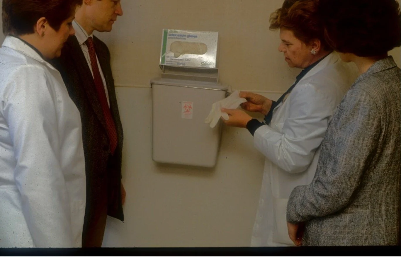 Four individuals, three women and one man, standing around a wall-mounted container for disposal of latex exam gloves, with leafy medical gloves on top, as one woman in a white lab coat shows a pair of gloves to the others.
