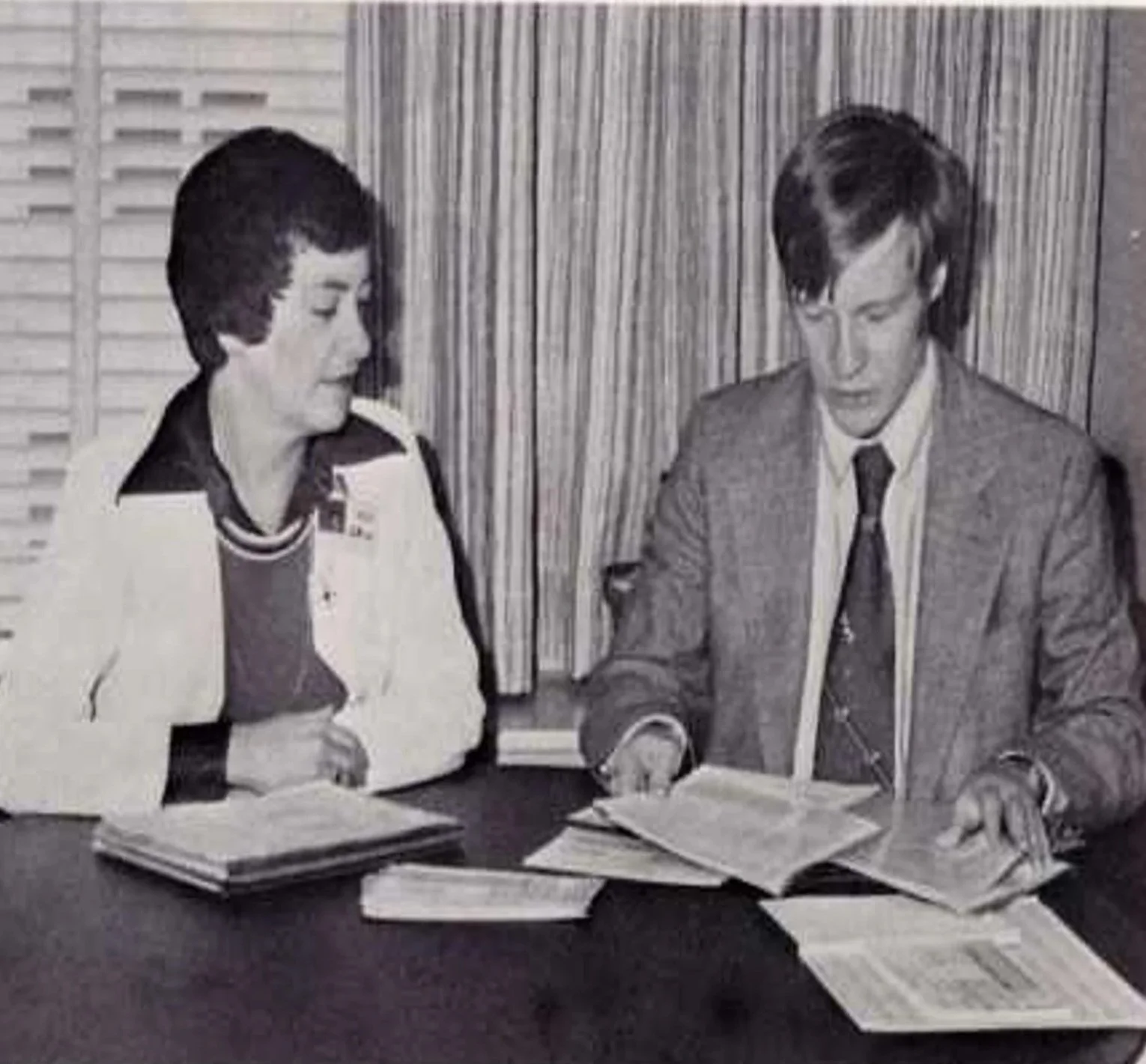 A woman and a man sitting at a table reviewing documents. The woman is wearing a sports jacket over a shirt, and the man is wearing a suit and tie. They are indoors with window blinds and a curtain in the background.