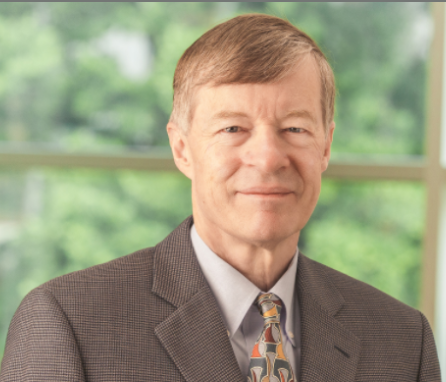 A middle-aged man in a gray suit and patterned tie, standing indoors in front of a window with greenery outside.