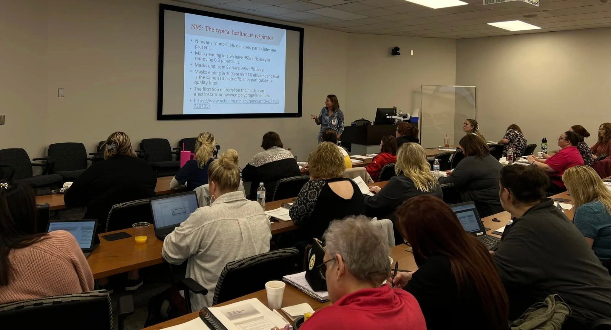 A woman is giving a presentation to a group of adults in a classroom or conference room. The presentation slide on the screen discusses the healthcare respirator N95.