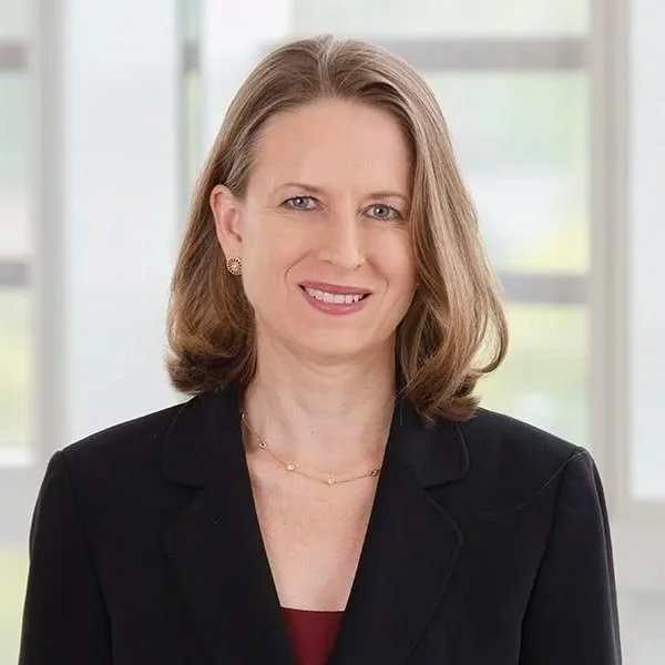 Portrait of a professional woman with light brown hair, wearing a black blazer and a red top, standing in front of a large window in a bright office setting.