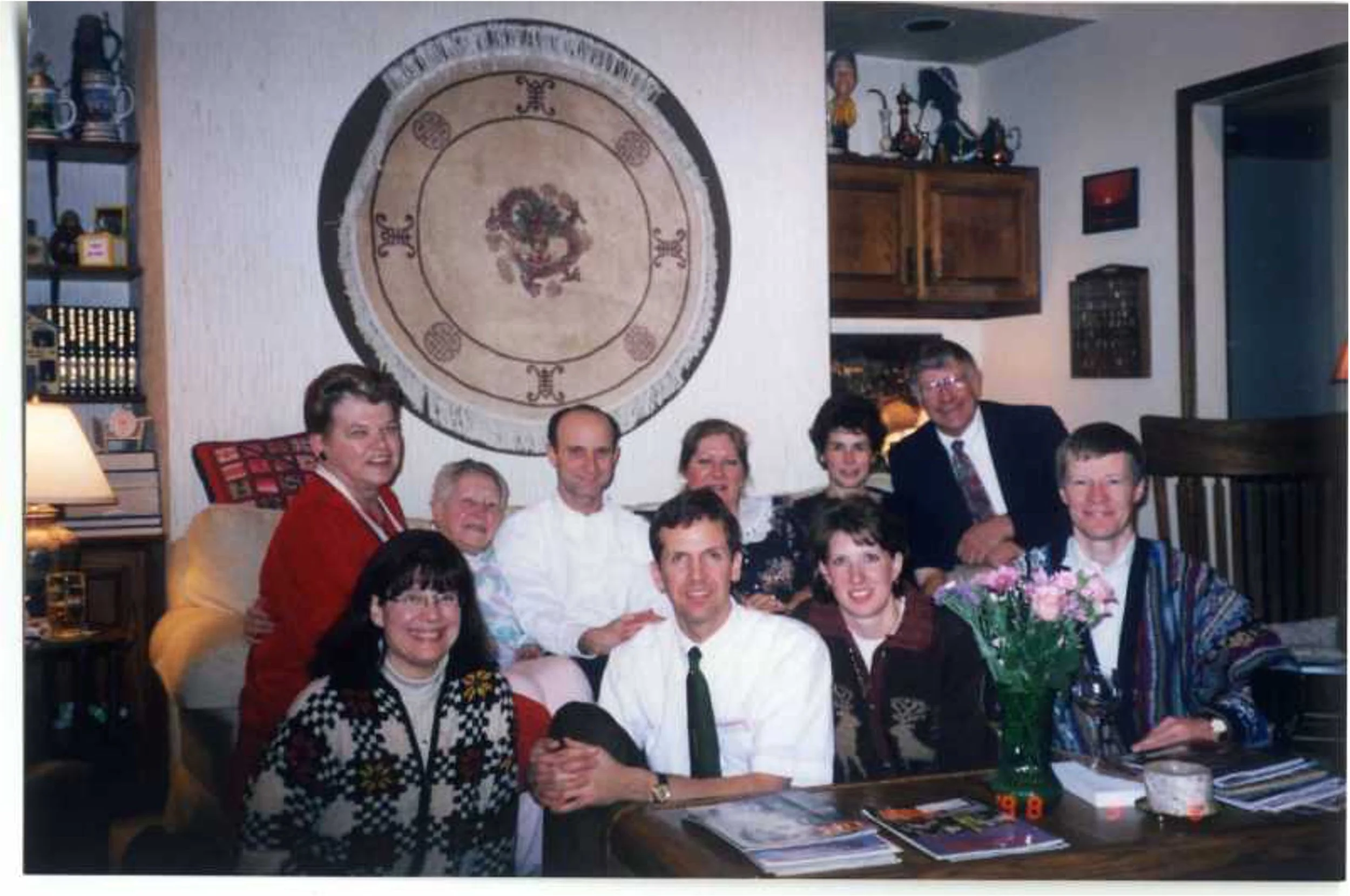 Group of ten people sitting and standing around a table with magazines and a vase of pink flowers in a cozy living room, smiling for the camera.