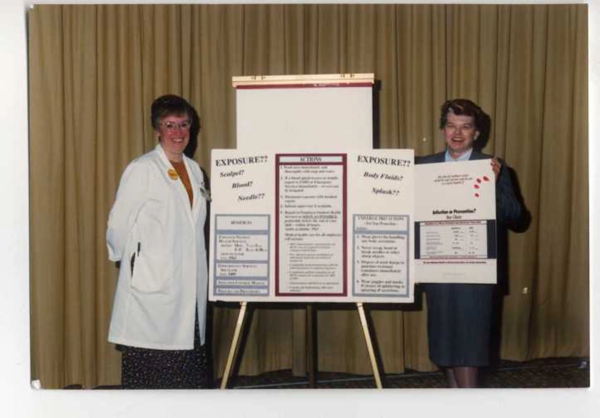 Two women standing beside a presentation board about exposure, with a yellow curtain in the background.
