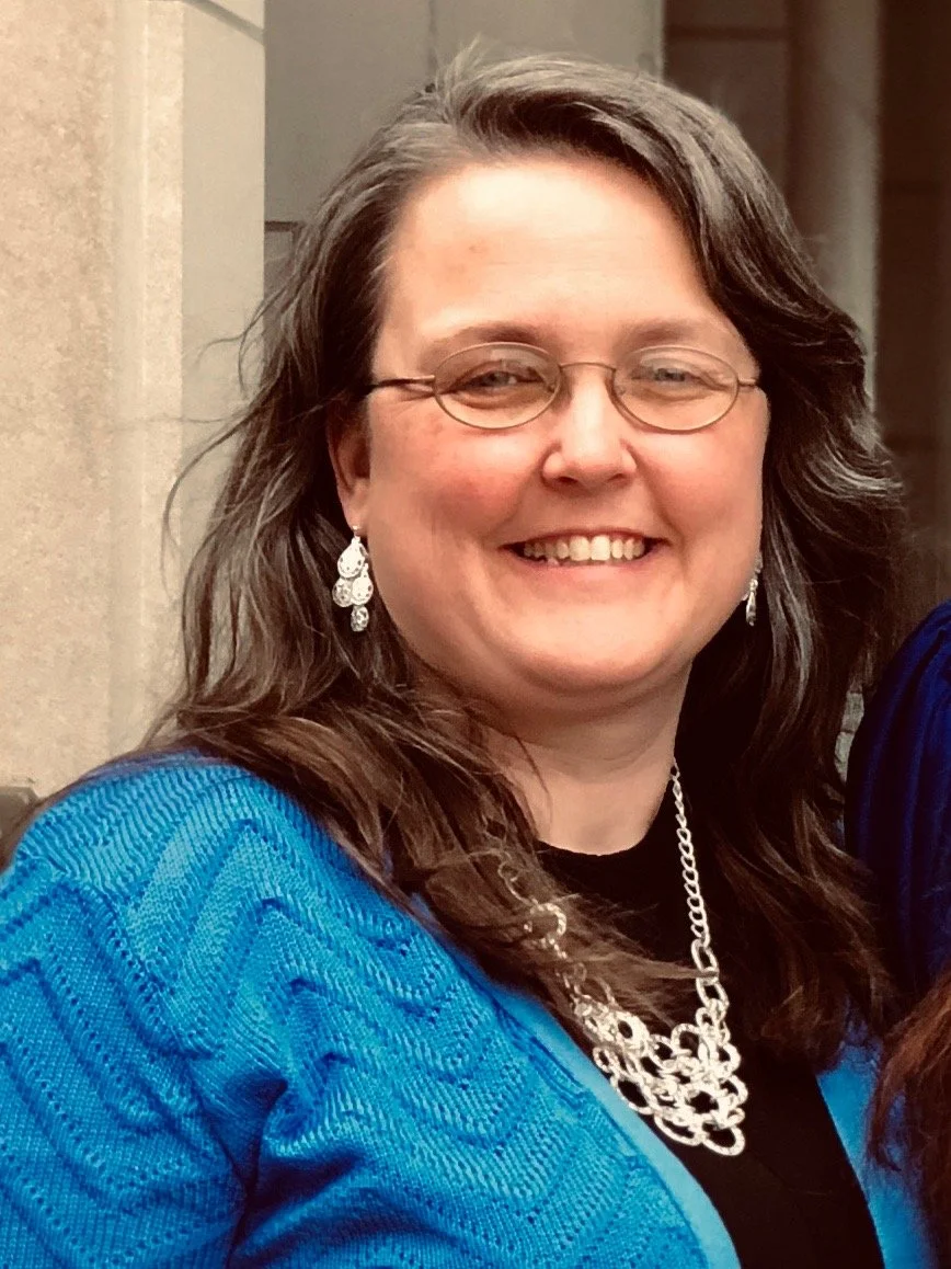 A woman with glasses, long brown hair, and light skin smiling, wearing a blue jacket, earrings, and a necklace.