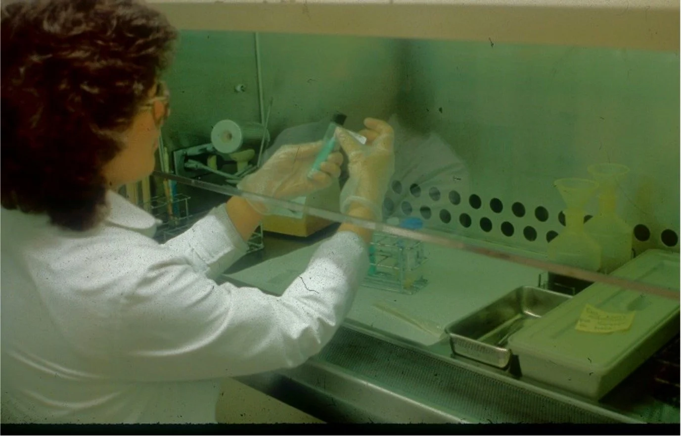 A scientist working in a laboratory hood, handling a test tube with a pipette, wearing safety gloves and glasses, surrounded by laboratory equipment and supplies.