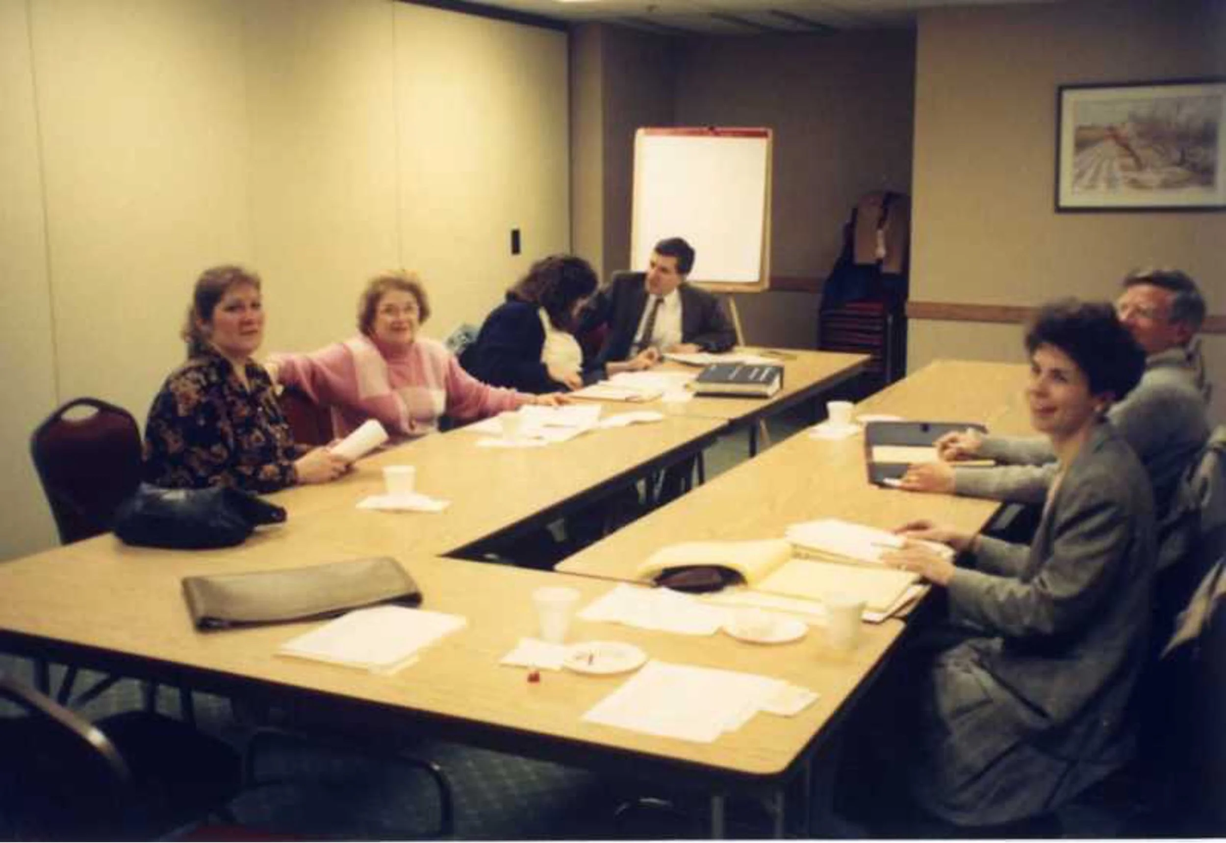 Group of six people sitting around a conference table in a meeting room, with papers, folders, cups, and a whiteboard behind them.