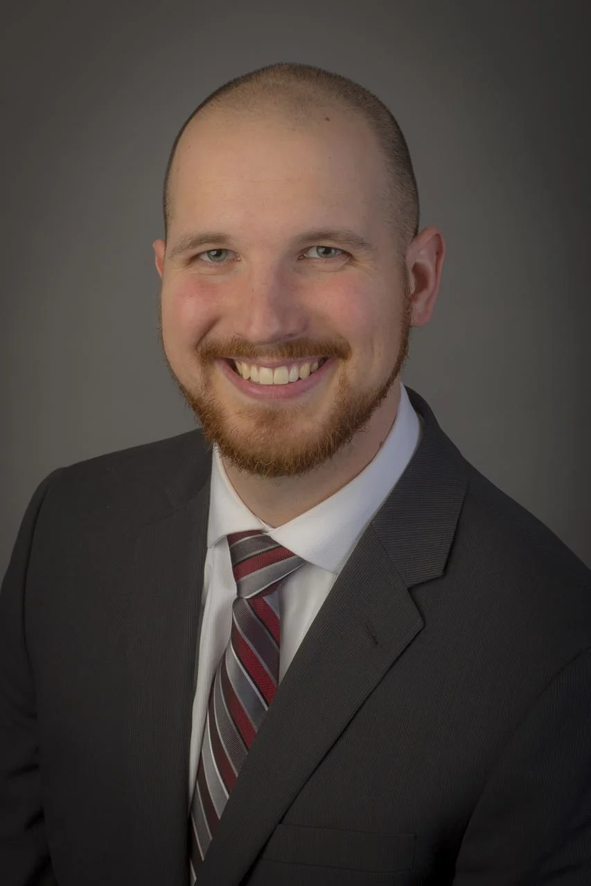 Headshot of a smiling man with a beard, wearing a dark suit and a striped tie, against a gray background.