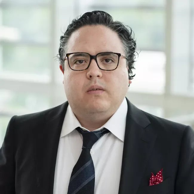 A man with dark, wavy hair and glasses wearing a dark suit, white shirt, and a striped tie.