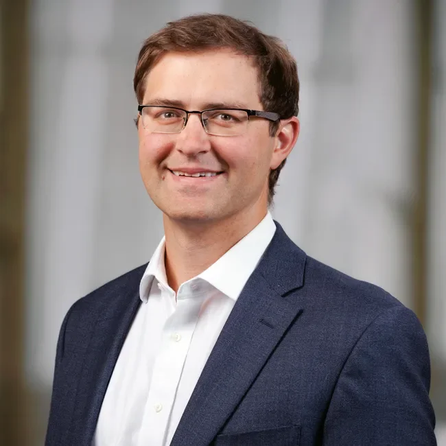 A professional man wearing glasses, a white shirt, and a navy blazer, smiling at the camera.