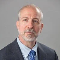 A professional headshot of a middle-aged man with gray hair and a beard, wearing a dark suit, light blue shirt, and blue tie, against a plain gray background.