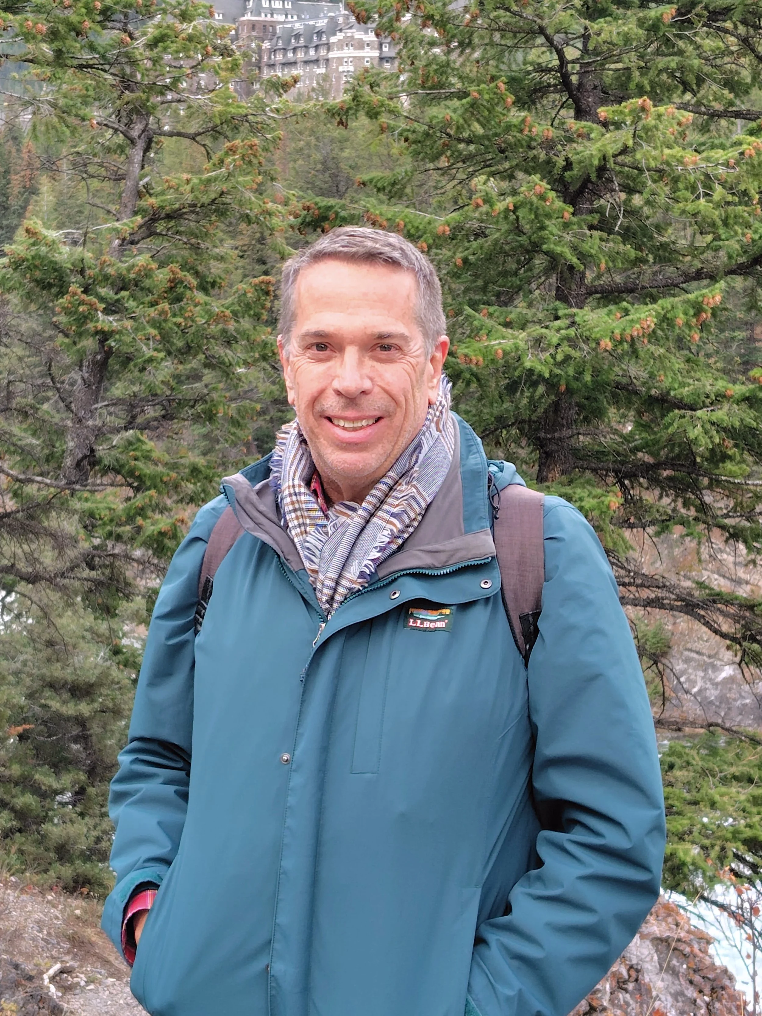 A smiling man in a teal jacket and scarf standing outdoors in a forested area with pine trees, with a mountain and a large hotel or building visible in the background.