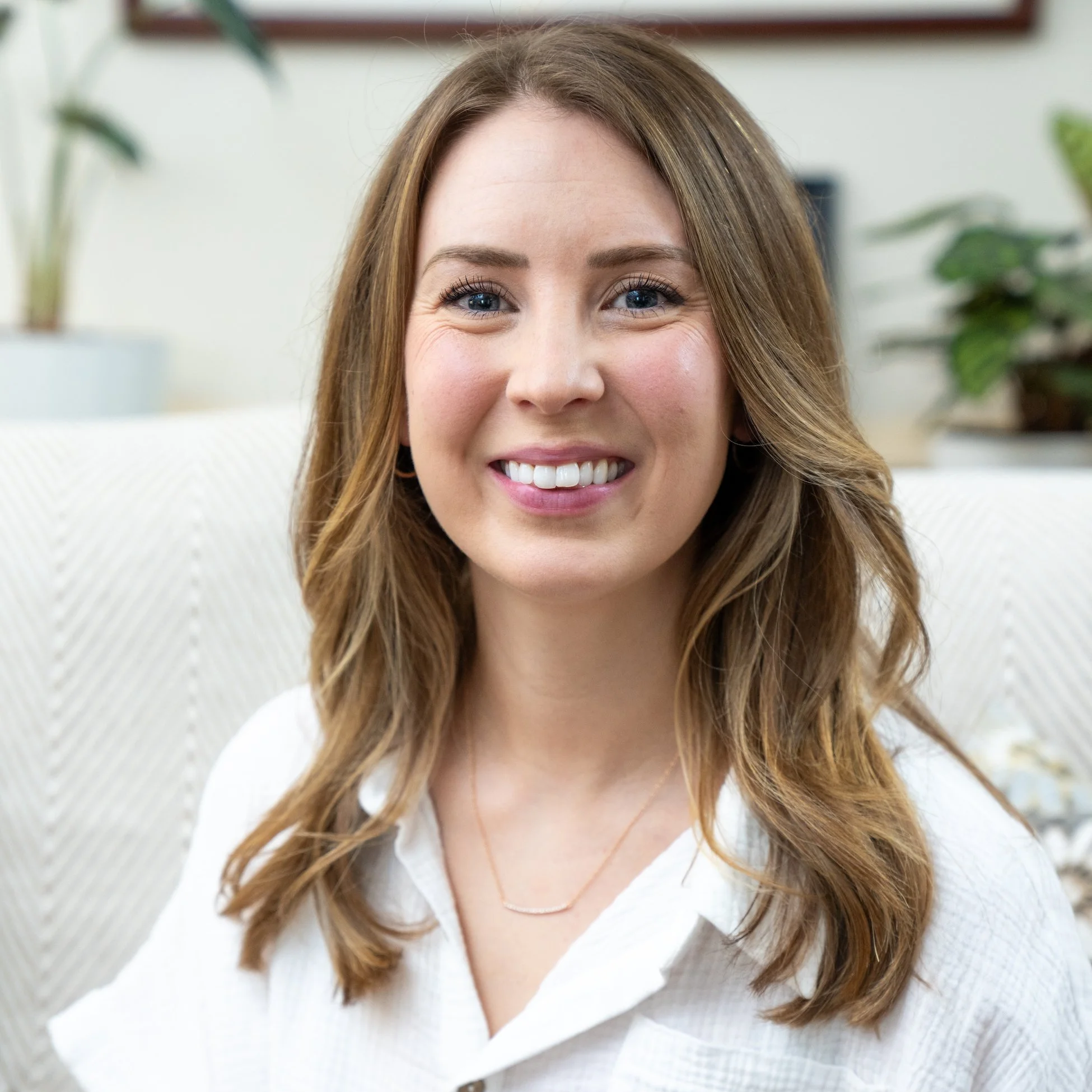 A smiling woman with long, wavy brown hair, wearing a white collared shirt and a delicate necklace, sitting in a cozy, well-lit room with plants in the background.
