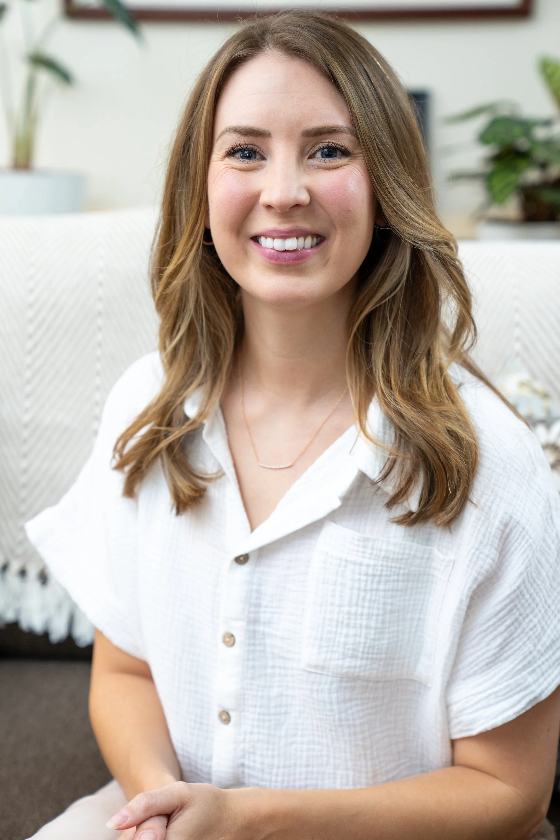 A young woman with wavy brown hair and blue eyes smiling at the camera, wearing a white button-up shirt and a delicate necklace, sitting indoors with houseplants in the background.