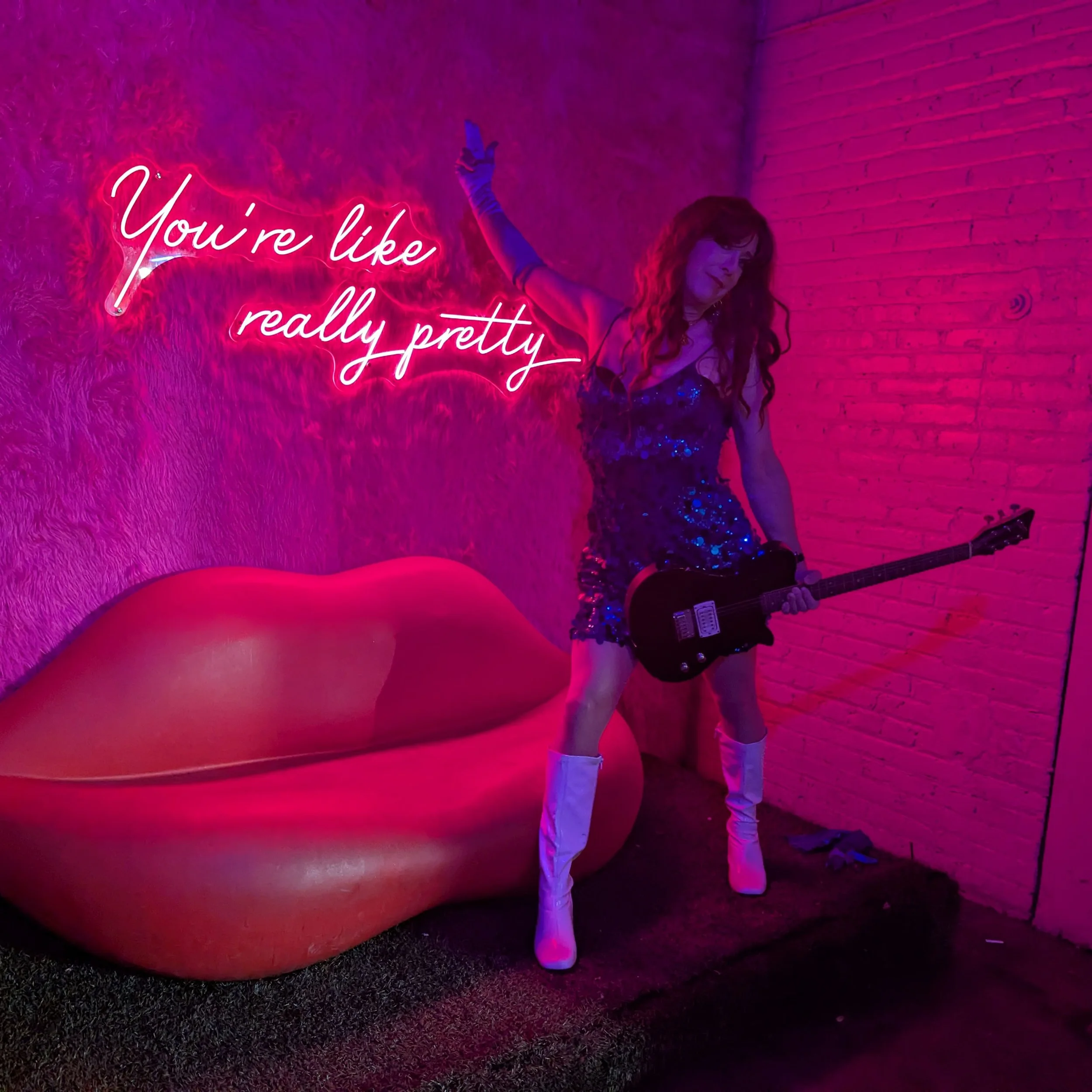 A woman stands next to a neon sign that says 'You're like really pretty,' holding a guitar and wearing a sparkling dress and white boots. The background features a pink and purple lighting with a lips-shaped chair nearby.