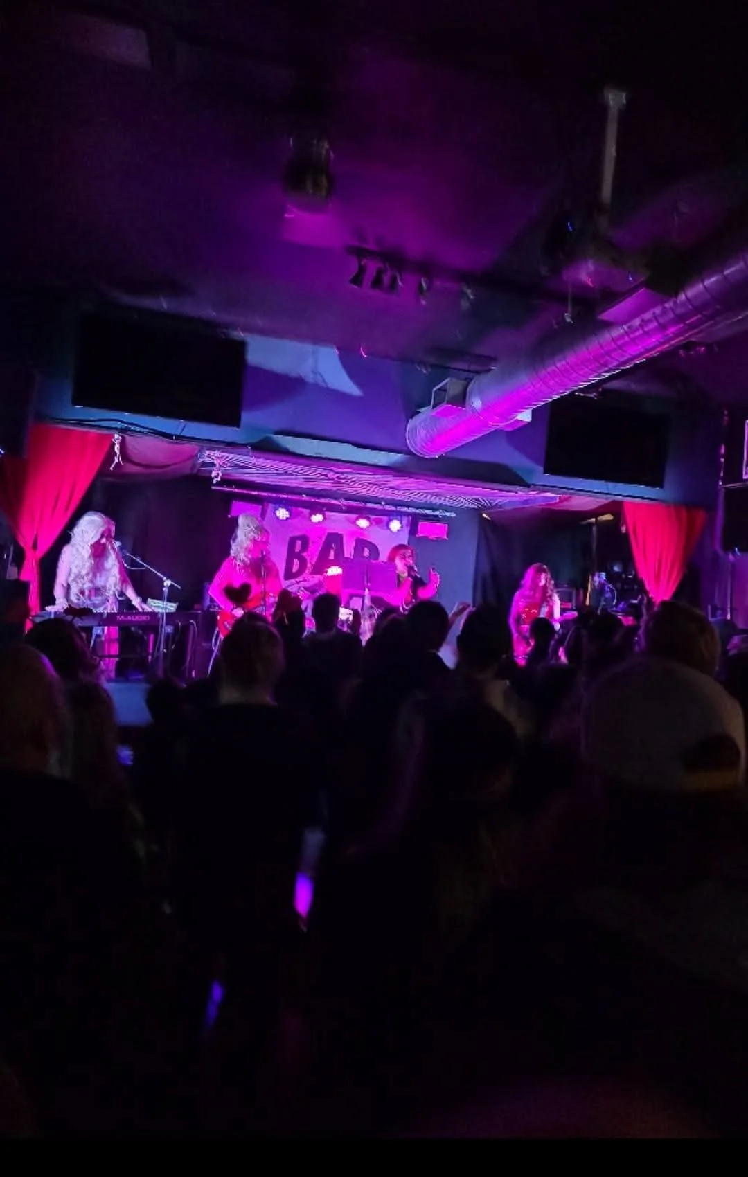 A live music performance on stage with a band playing, colorful purple lighting, and an audience watching.