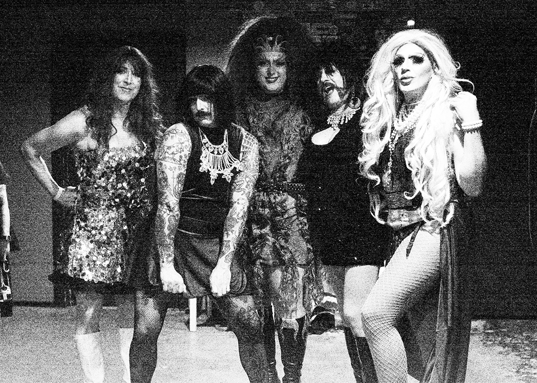 Black and white band photo of five women dressed in elaborate, glamorous drag costumes and makeup, standing together in a line.