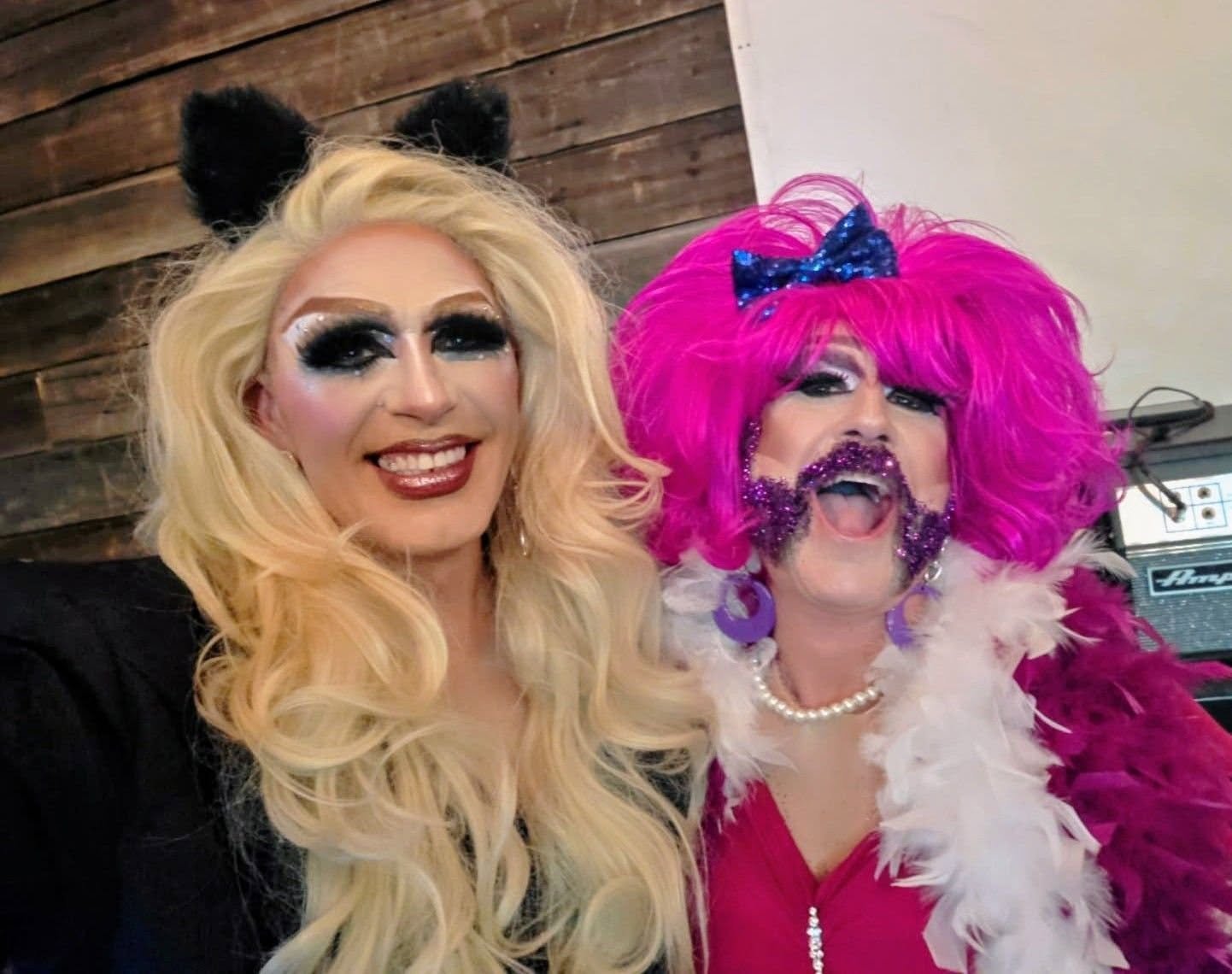 Two drag queens posing together; one with blonde hair, panda ears, and black makeup, and the other with pink hair, glittery makeup, purple earrings, and feathered costume.