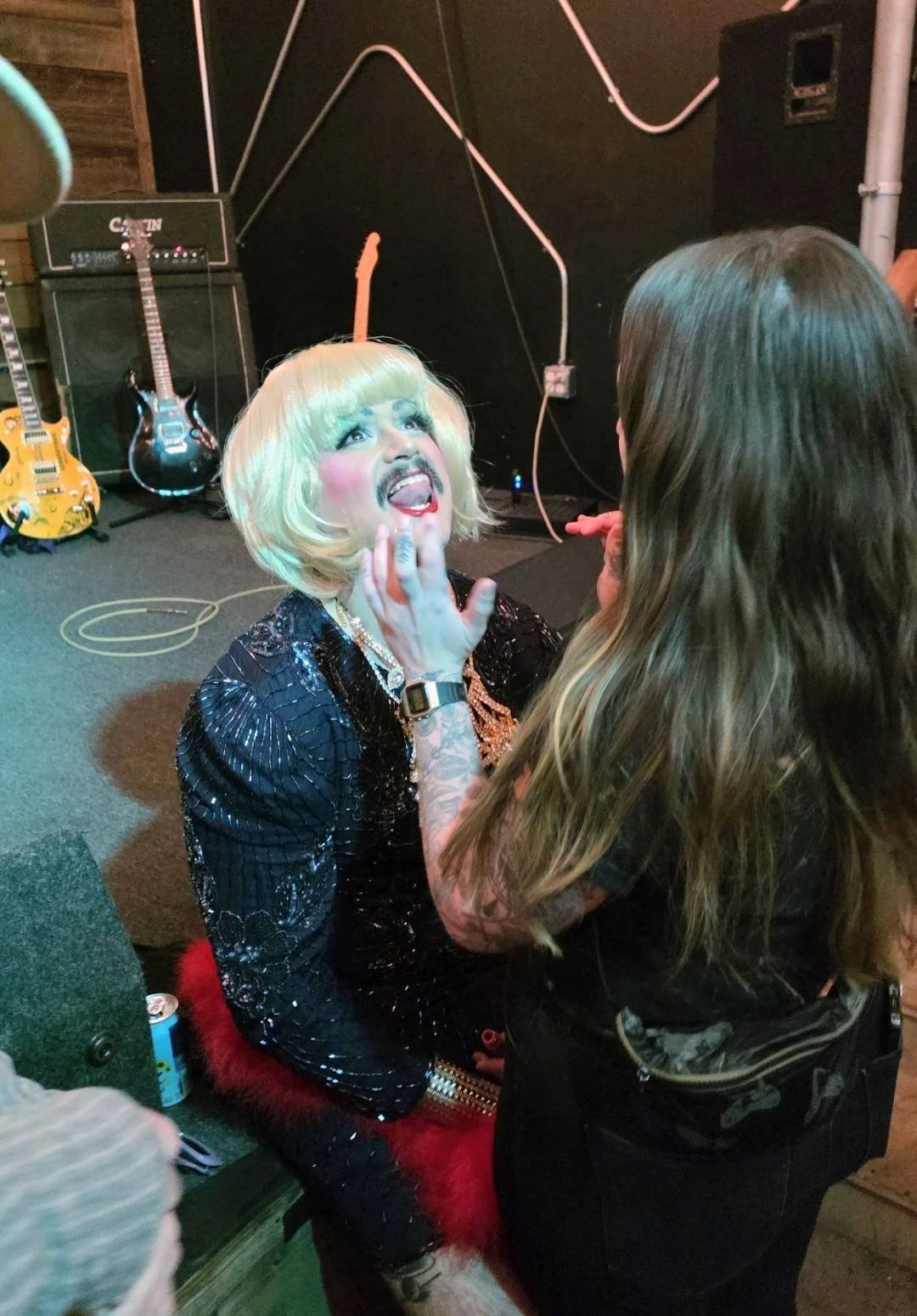 A person with blonde wig, makeup, and sequined black outfit sits on a chair, holding their face with their hand while talking to a woman with long brown hair in a black top. The scene is in a music venue with guitars and amplifiers in the background.